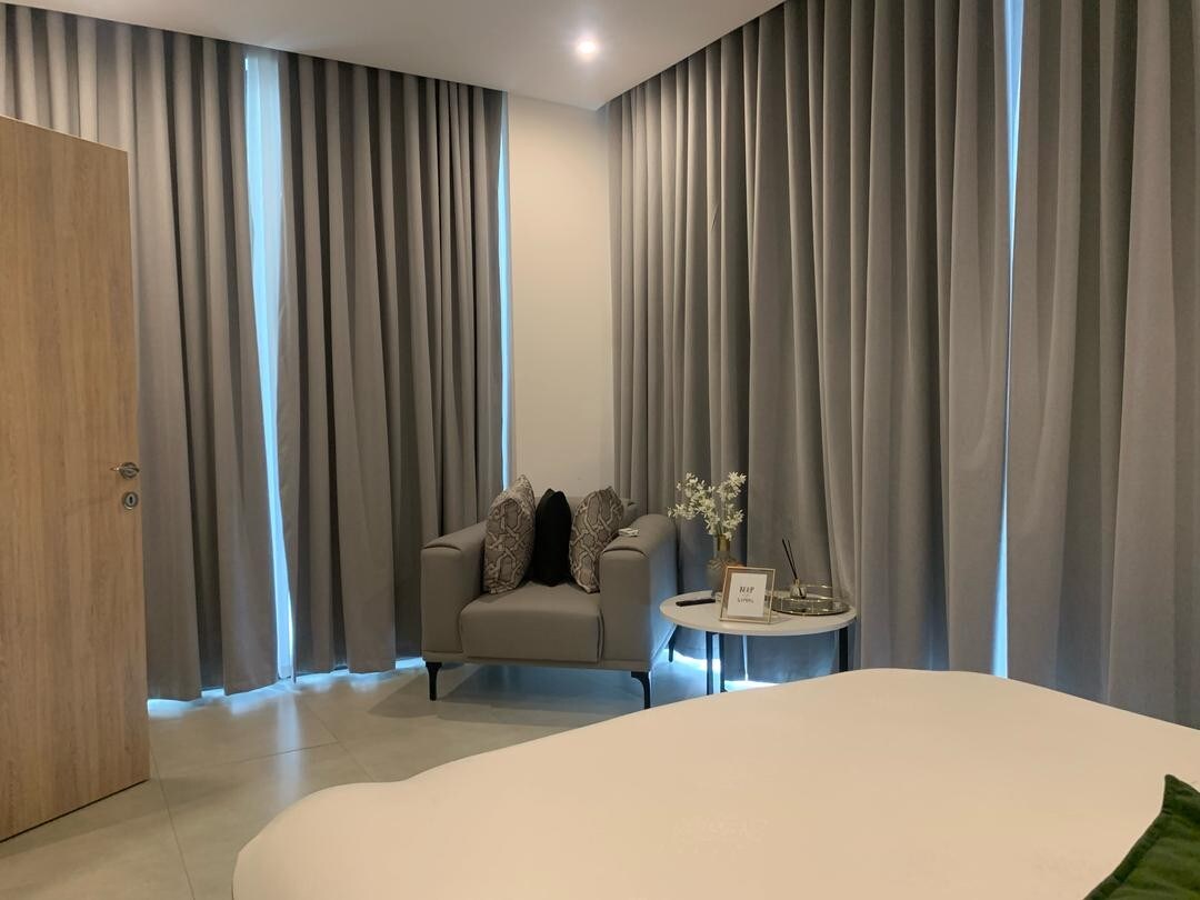 A sleeping area is complemented by soft, gray curtains that frame a large window. A modern armchair with decorative pillows is positioned beside a small accent table. Natural light is gently filtered through the curtains, creating a serene atmosphere.