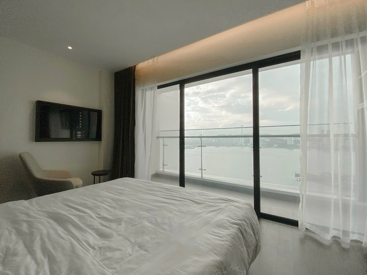 A spacious room features a large bed with a neatly arranged white comforter. Floor-to-ceiling windows offer unobstructed views of the water, with sheer curtains softly filtering natural light. A comfortable chair and a television are positioned against the wall.