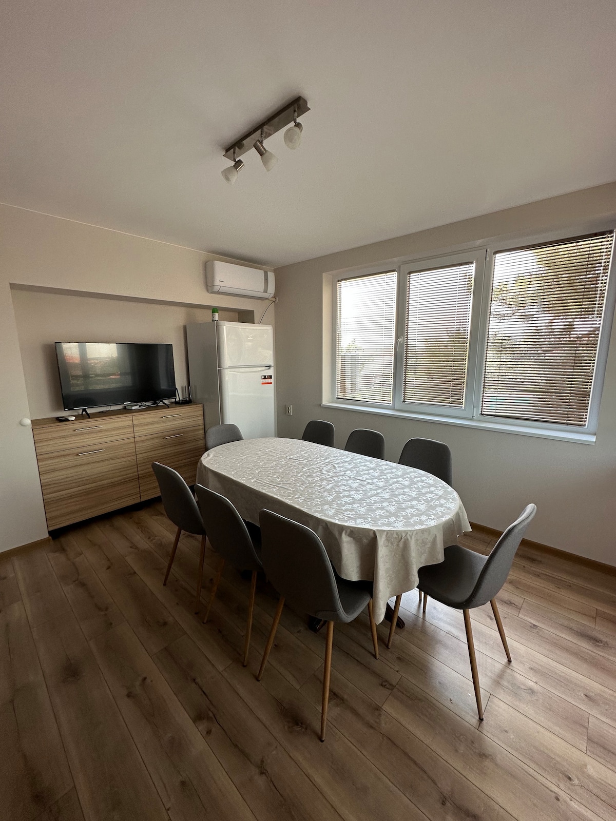 A dining area is presented, featuring a large oval table surrounded by six chairs. Natural light enters through three windows, creating a bright atmosphere. A television and a refrigerator are positioned against a wooden cabinet, while air conditioning is visible on the ceiling.