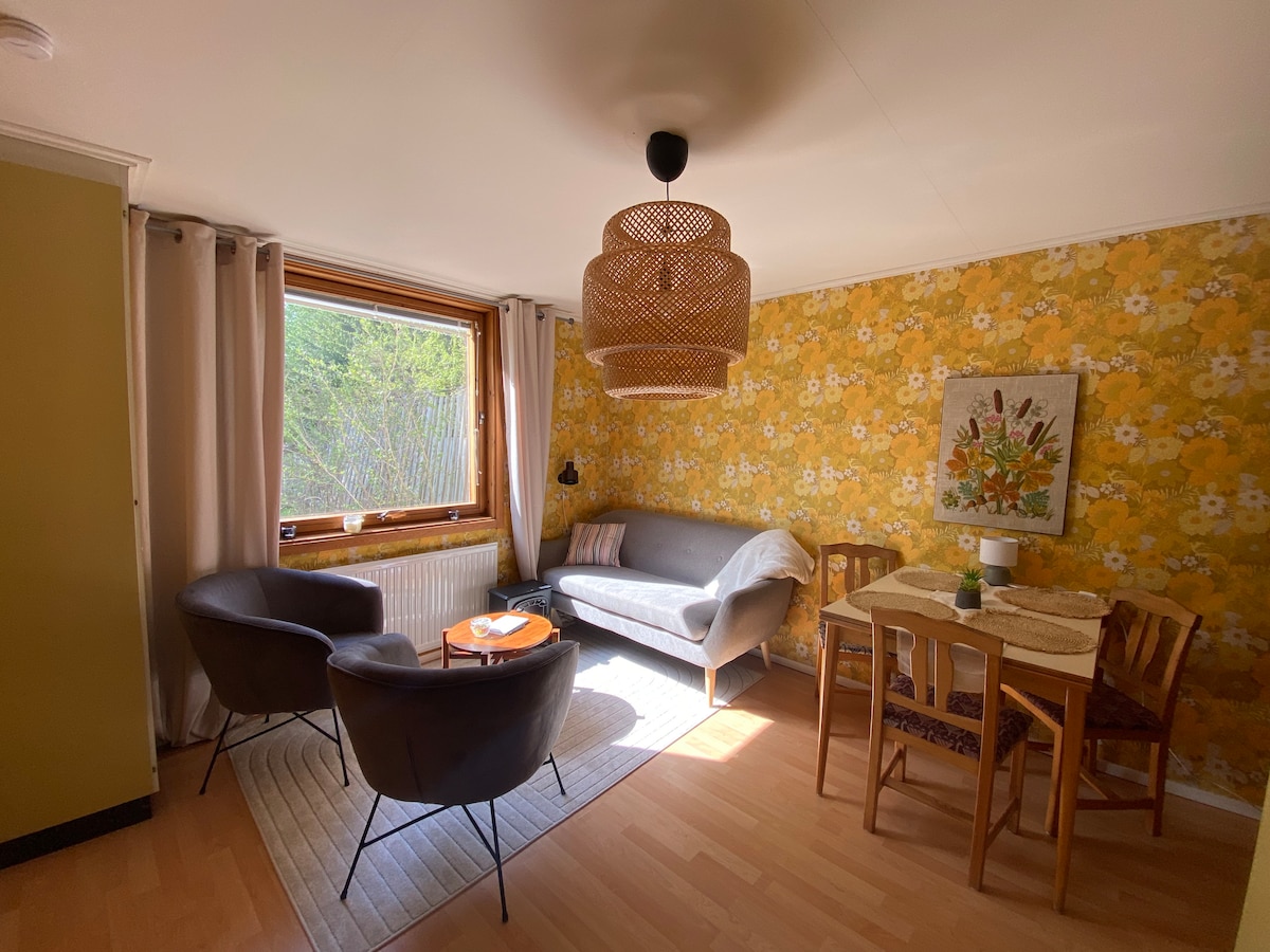 A bright living area features a floral wallpaper and a mix of modern and vintage furnishings. A cozy sofa and two armchairs surround a small coffee table, while a dining table is set for two. Natural light filters in through a large window, enhancing the warm atmosphere.