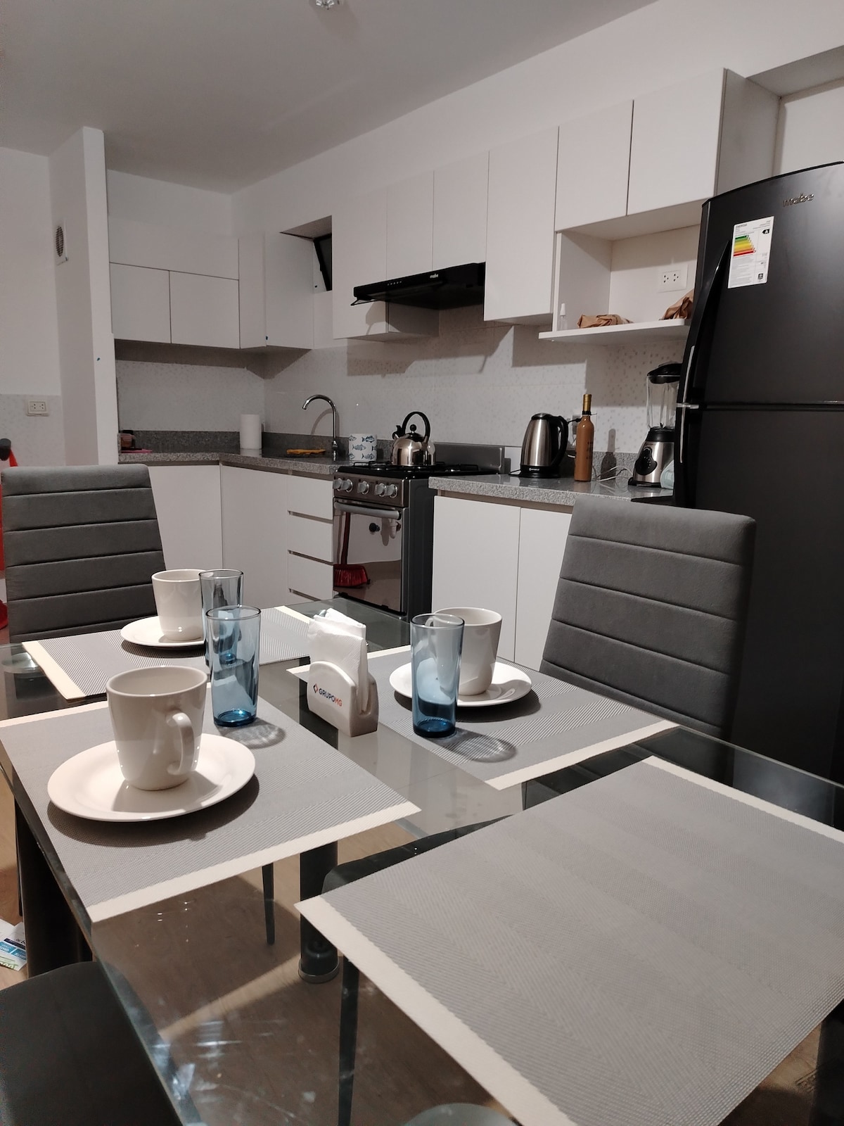 A well-equipped kitchen is visible, featuring modern appliances including a stovetop, kettle, and refrigerator. A dining table is set for four, with plates, glasses, and utensils arranged neatly. Light-colored cabinets and a granite countertop complement the spacious and organized layout.