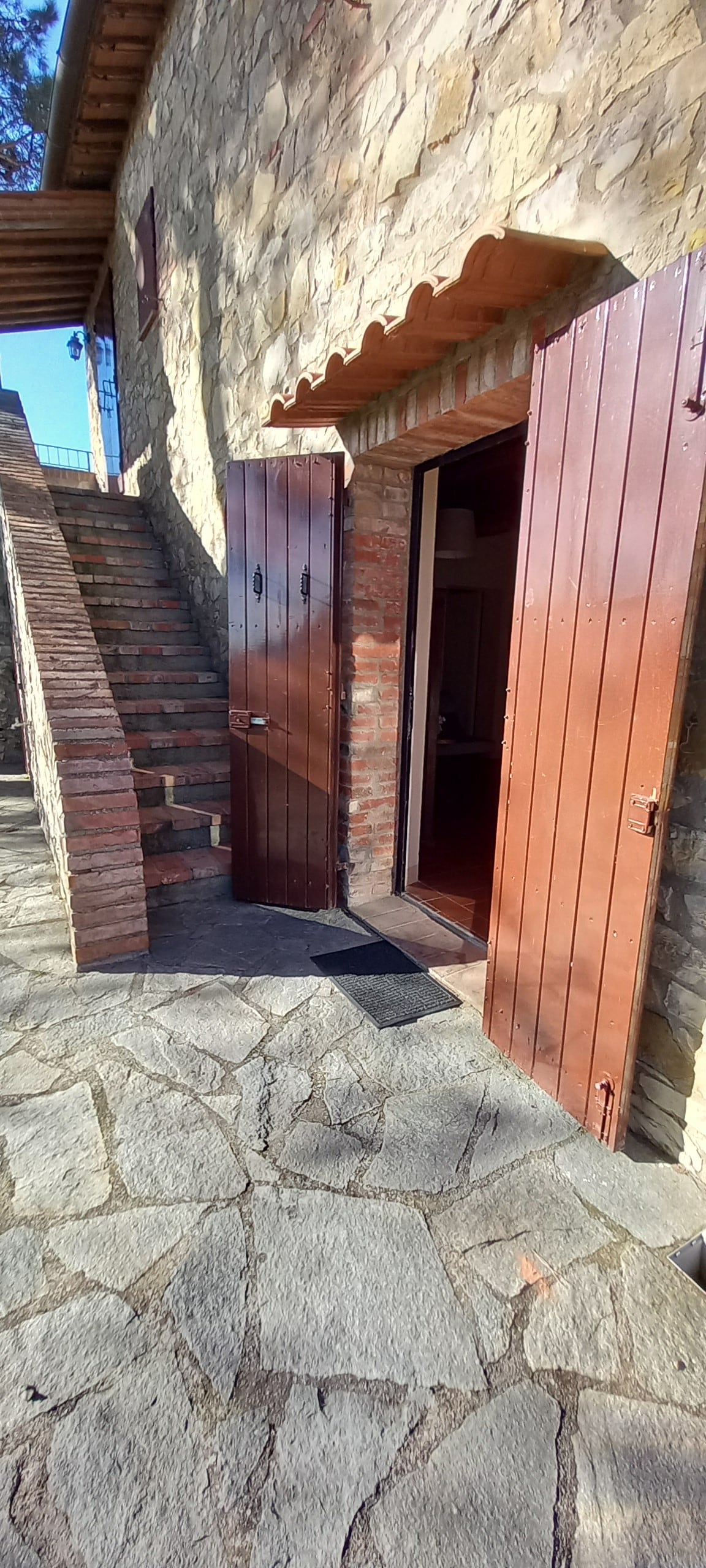 A rustic stone exterior showcases an entrance with double wooden doors that open to a welcoming space. Natural stone flooring leads the way, and a set of stairs ascends to the upper level, framed by warm afternoon light illuminating the surroundings.