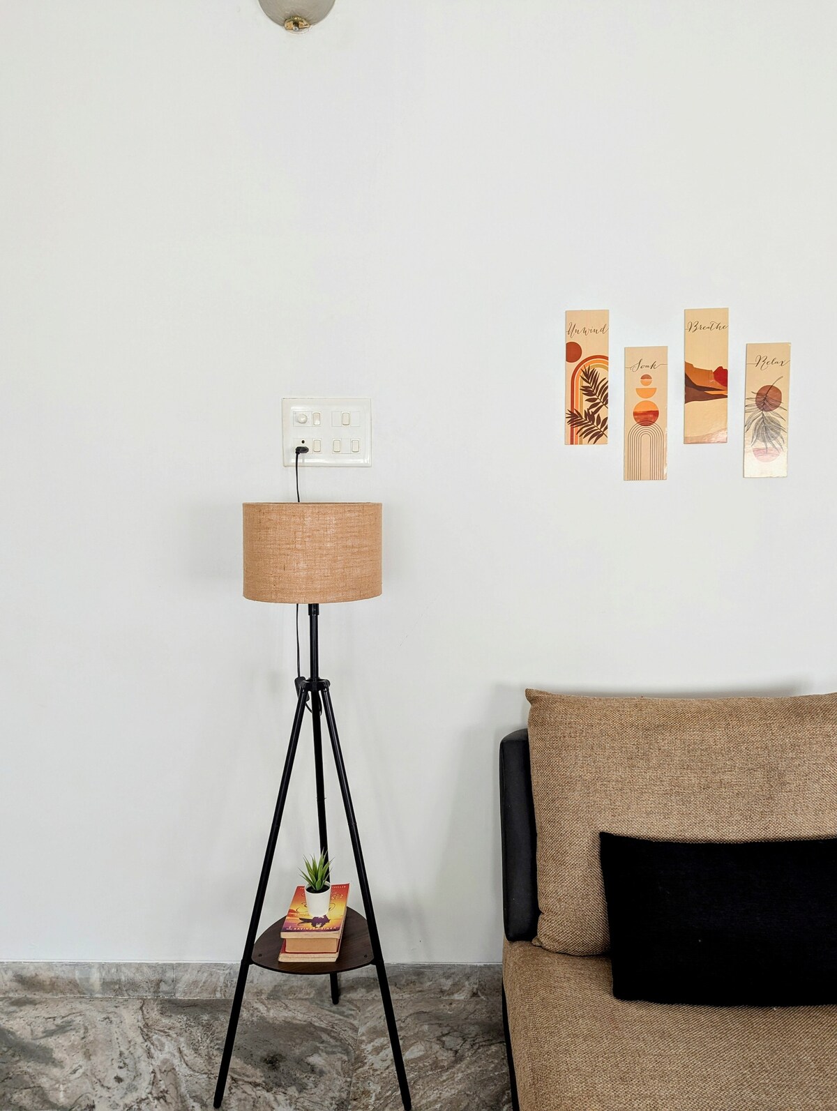 A neutral-colored wall features three decorative art pieces, each displaying abstract designs and earthy tones. A stylish floor lamp with a beige shade stands beside a brown sofa, while a small potted plant rests on a stack of books beneath the lamp, adding a subtle touch of greenery.