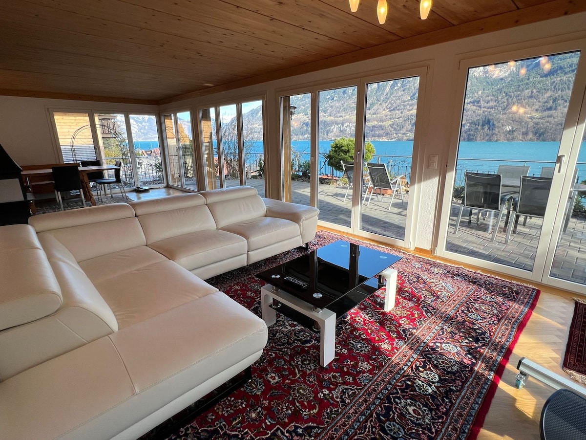 A spacious living area features a comfortable white sofa set and a low black coffee table atop a patterned rug. Expansive glass doors frame views of Lake Brienz and the surrounding mountains, filling the space with natural light. A dining table is positioned to the side.