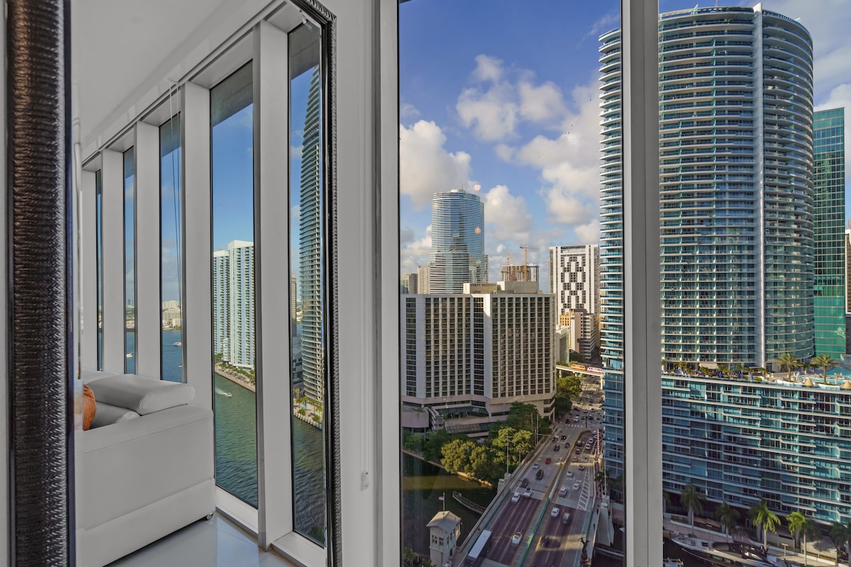 Floor-to-ceiling windows reveal panoramic views of the ocean and Miami skyline. The scene captures a bright and airy atmosphere, with soft natural light flooding the space. The sleek furniture complements the modern design, enhancing the spacious feel of the corner unit.