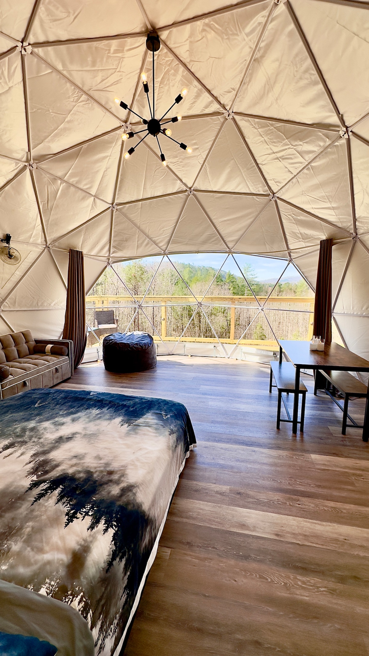 The interior of the geodesic dome features a queen bed with a scenic bedspread, a cozy sofa, and a bean bag chair. A dining table with seating is positioned near the glass windows, offering views of the surrounding nature. Natural light illuminates the space through the dome's triangular design.