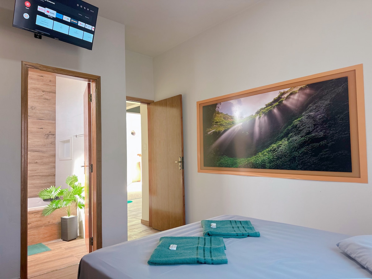 A well-lit room features a spacious bed neatly made with towels arranged at the foot. A large wall mural depicting a serene natural landscape adds a calming touch. The entrance to a bathroom is visible in the background, enhancing the room's accessibility.