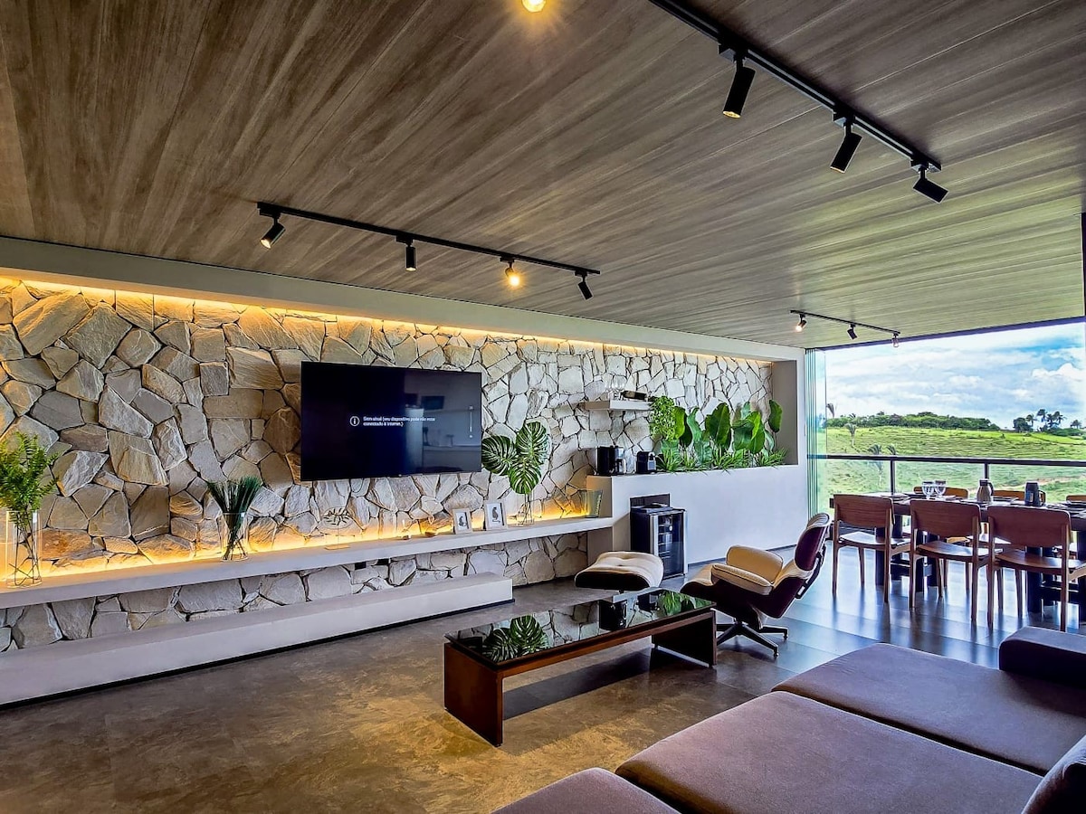 A modern living area is characterized by a stone accent wall and natural lighting. A contemporary sofa is positioned in front of a sleek entertainment unit featuring a television. The space opens to an outdoor dining area with a view of the surrounding landscape.