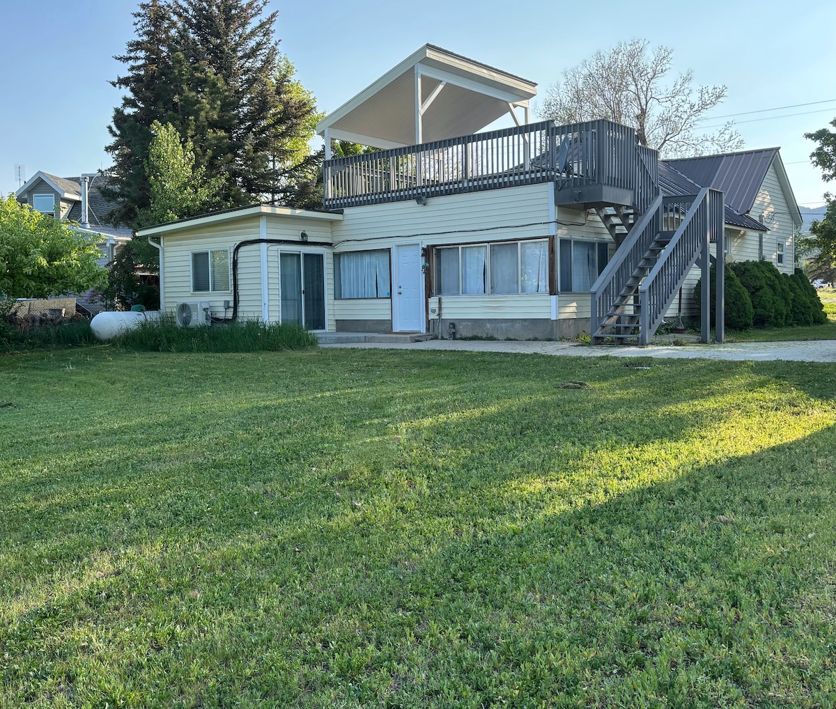 A two-story house is set on a large green lawn, featuring a spacious upper deck and a staircase leading to it. The exterior consists of light-colored siding, and multiple windows reflect natural light. Surrounding foliage adds to the peaceful outdoor environment.