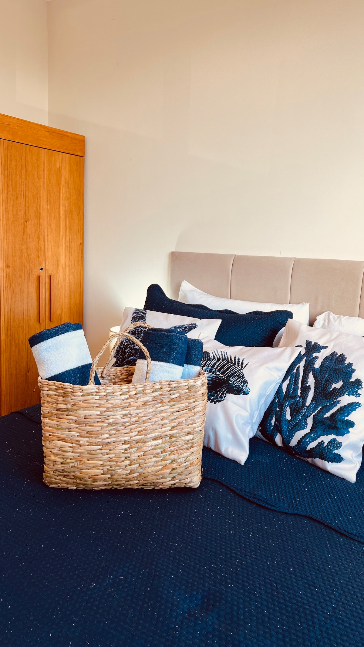A neatly made bed features a navy blue blanket and contrasting white pillows adorned with decorative patterns. A woven basket containing towels rests at the foot of the bed, while a wooden wardrobe stands beside it, contributing to a clean and organized look.