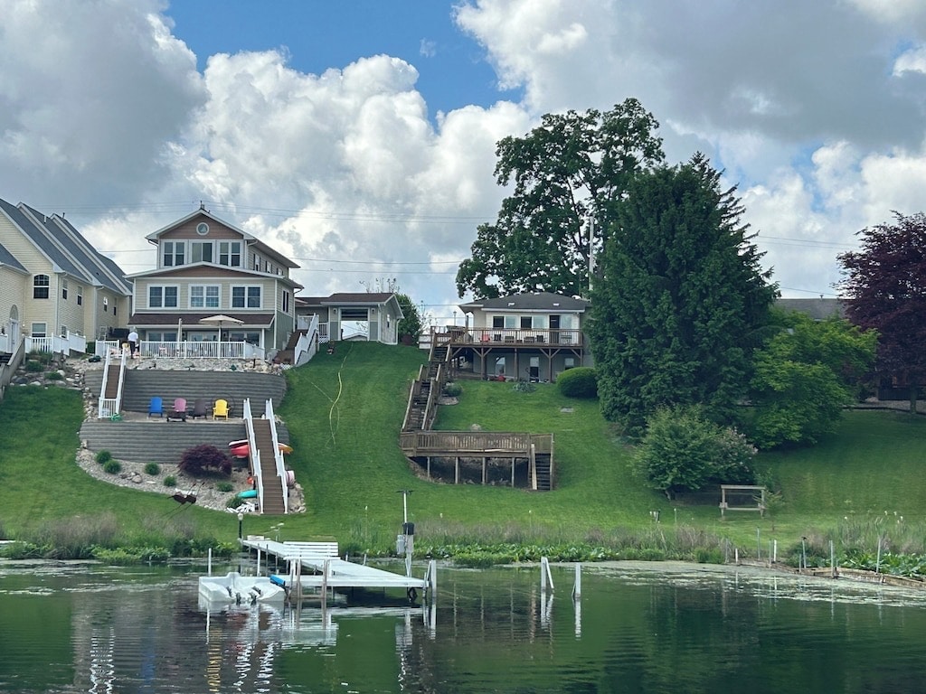A tranquil lakeside view showcases a grassy slope leading to three multi-level homes. Colorful chairs are placed along the upper deck, while a wooden dock extends towards the calm water. Lush trees and shrubs enhance the natural setting.