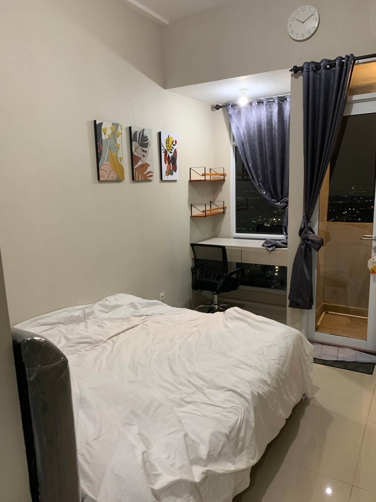 A well-lit sleeping area features a spacious bed with a white coverlet. Above the bed, three colorful artwork pieces are displayed on the light grey wall. A small desk and chair are positioned by the window, which is adorned with a dark curtain, revealing a nighttime city view.