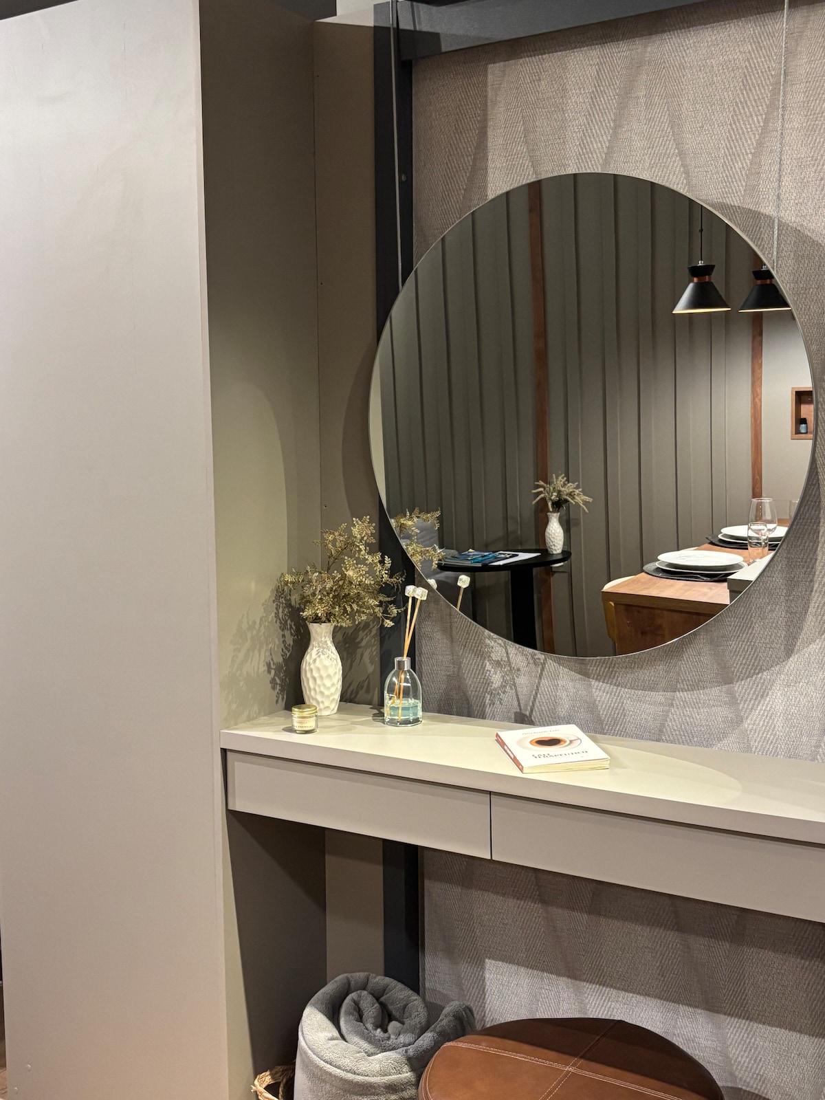 A modern console table is displayed against a textured wall, featuring a round mirror that reflects the room. A vase with dried flowers and a decorative bottle are positioned on the table. Soft towels and a small leather ottoman are neatly arranged below.