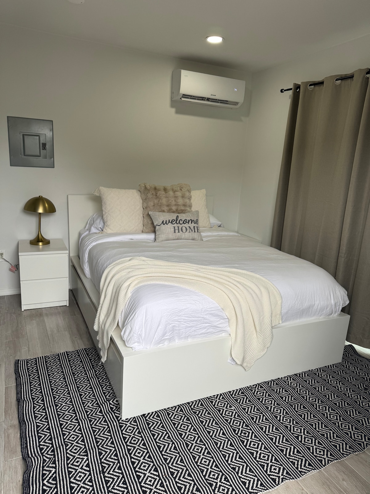 A well-appointed sleeping area features a spacious bed with white linens and decorative pillows. A soft throw blanket is draped over the bed. A contemporary nightstand with a lamp stands beside the bed, and light enters through the partially drawn curtains.