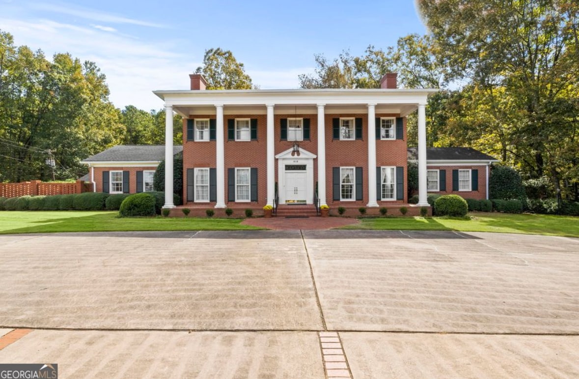 A grand two-story Southern estate is showcased, featuring a classic facade with white columns and a brick exterior. Lush greenery surrounds the property, enhancing its elegance. A spacious driveway leads to the welcoming entrance, framed by well-maintained landscaping and symmetrical windows.