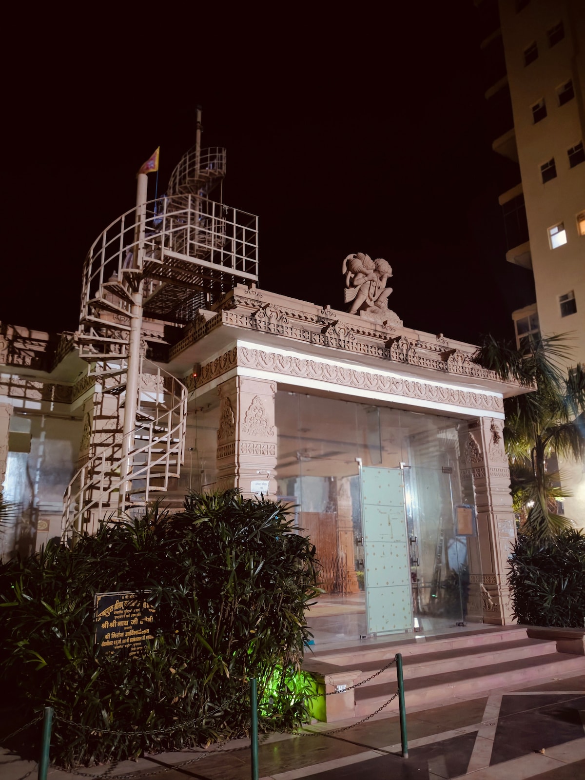 The entrance of a beautifully crafted temple is illuminated at night, showcasing intricate stone carvings. Lush greenery surrounds the structure, with a staircase beside it leading to an upper level. Soft lighting enhances the temple's details, creating a serene ambiance.