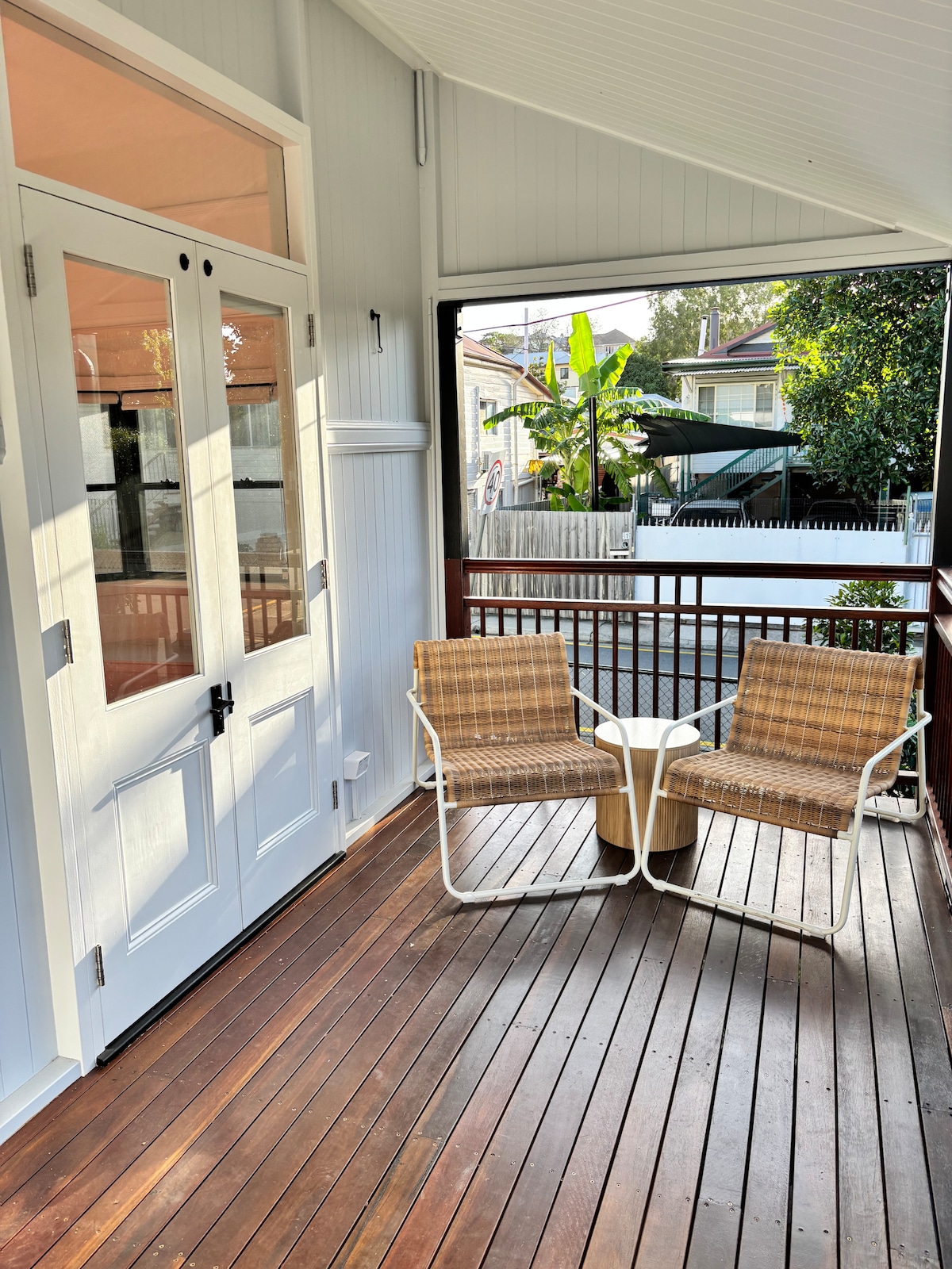 A covered porch area features two outdoor chairs positioned around a small table. Natural light filters in through the French doors, and the wooden flooring provides a warm tone. The surroundings include glimpses of greenery and nearby houses.