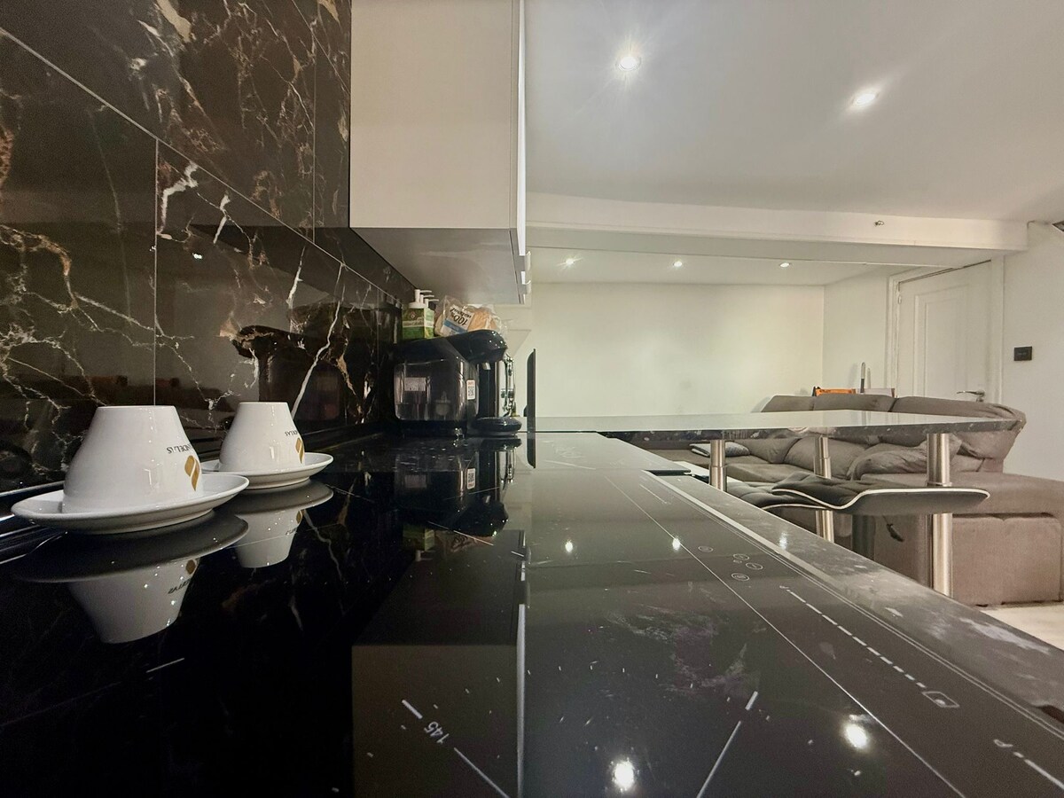 A modern kitchen space is highlighted, featuring a sleek black countertop and a coffee maker. White cups are set on the counter, and a cozy living area is visible in the background, furnished with a light-colored couch.