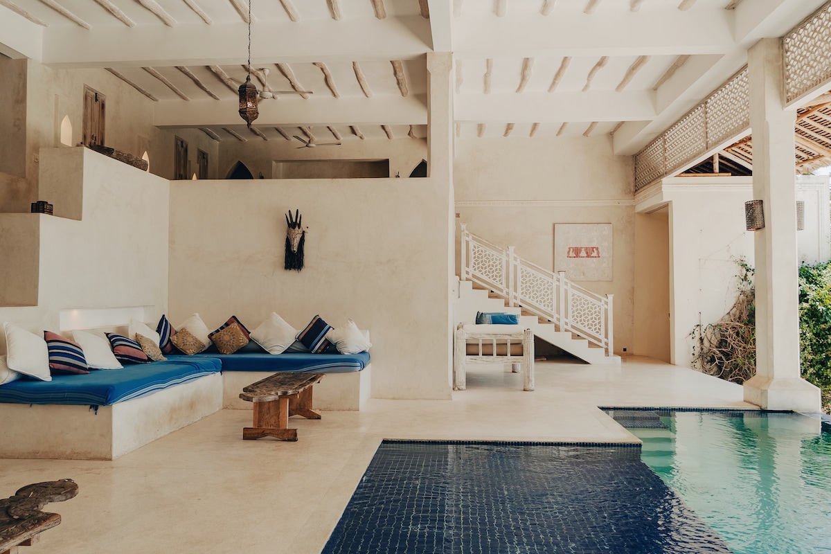An indoor area showcases a serene space with soft seating arranged along one wall, accented by colorful cushions. A staircase leads to the upper level, while a swimming pool is visible to the right. Natural light floods the space, enhancing the soothing ambiance.
