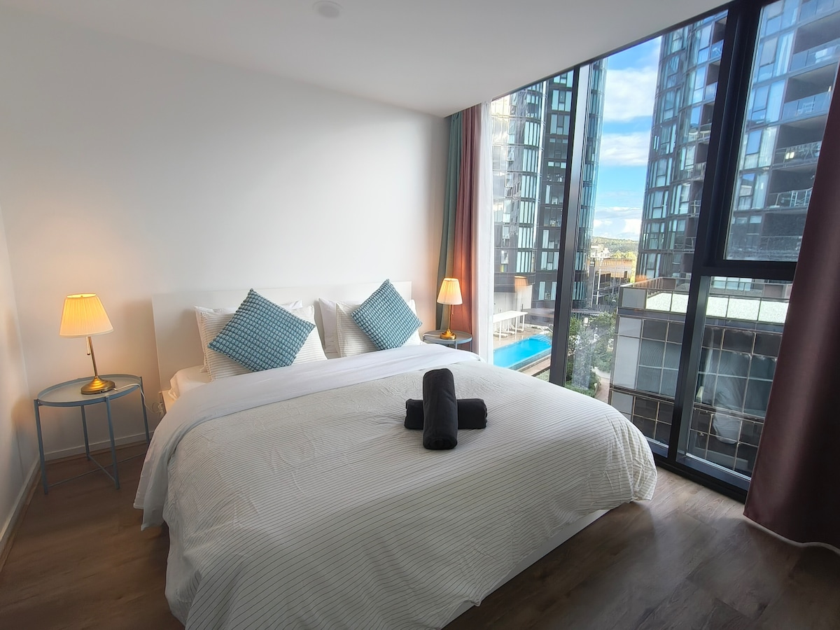 A well-lit bedroom is furnished with a spacious bed dressed in crisp white linens, accented by two decorative pillows. A bedside table holds a lamp, providing gentle illumination. Large windows allow natural light to fill the room, offering views of the surrounding cityscape.