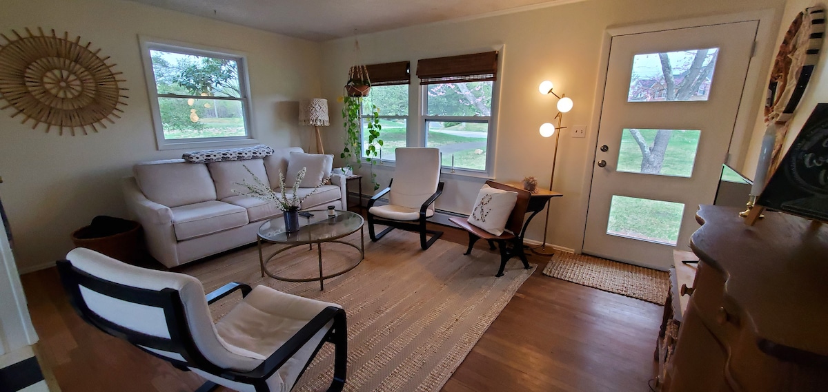 A welcoming living room is presented, featuring a light-colored sofa, two armchairs, and a glass coffee table. Natural light enters through the windows, illuminating the space. A decorative wall piece is displayed, alongside greenery from potted plants, enhancing the inviting ambiance.