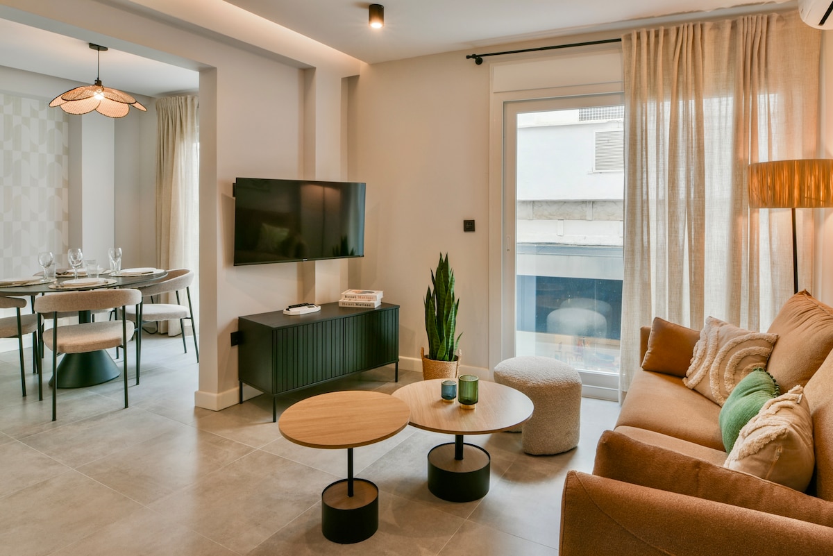 A modern living area is presented with a comfortable sofa and stylish coffee tables. Natural light filters through sheer curtains, illuminating the space. A flat-screen TV is mounted on the wall, while a potted plant adds a refreshing touch.