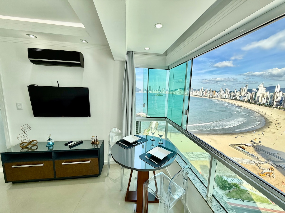 A modern dining area features a round table set for two, positioned next to expansive glass windows that frame a panoramic view of the beach and skyline. A wall-mounted television is visible, complemented by a sleek media console. Natural light fills the space.