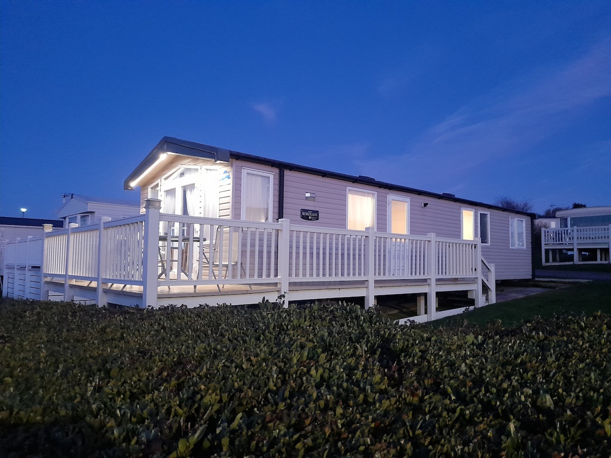 A holiday home is depicted at dusk, featuring a spacious wrap-around deck. The exterior walls are beige, with multiple windows reflecting the evening light. A low hedge surrounds the area, providing privacy and greenery.