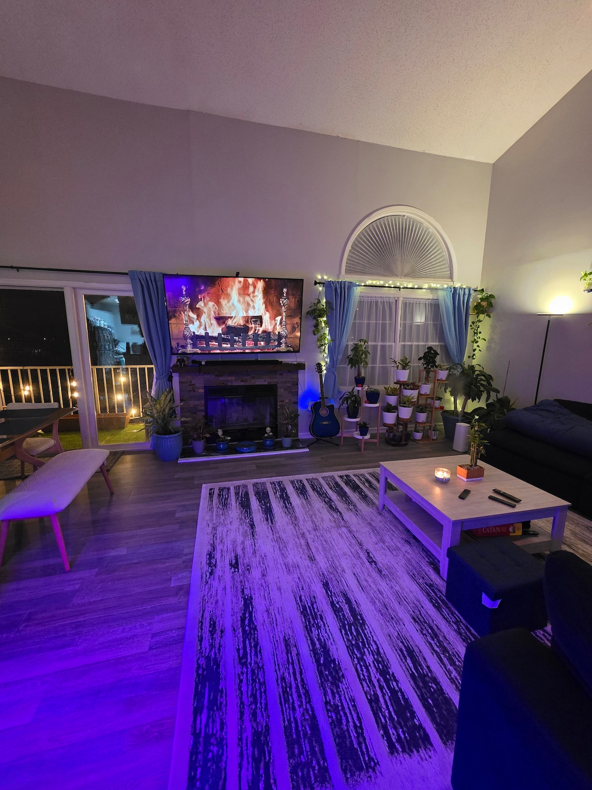 A cozy living room features a stylish fireplace below a large television displaying a fire simulation. Soft lighting enhances the room's atmosphere, with plants adorning the space. A comfortable sofa and a coffee table complete the inviting layout, with a balcony visible through glass doors.