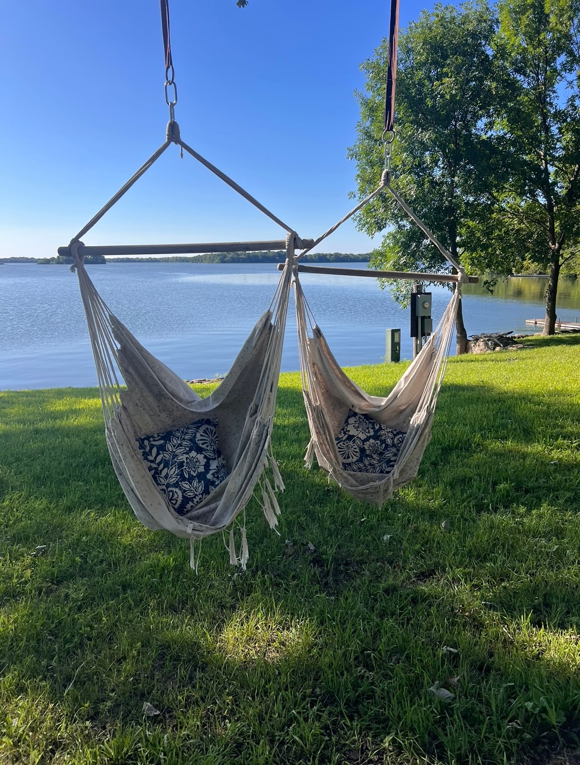 Two hanging chairs are positioned near the water's edge, offering a serene view of the calm lake. The chairs feature patterned cushions, and a lush green lawn surrounds them. Tall trees provide shade and enhance the peaceful atmosphere.