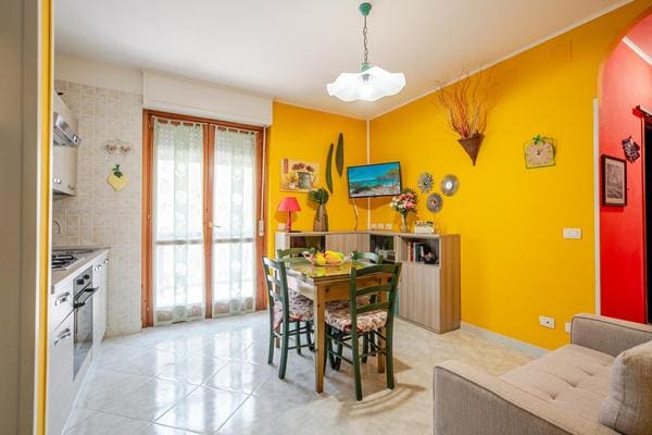 A bright kitchen area features a dining table with chairs, complemented by a small TV mounted on the wall. The walls are painted a warm yellow, enhancing the inviting atmosphere. Natural light enters through the sliding doors leading to a balcony.