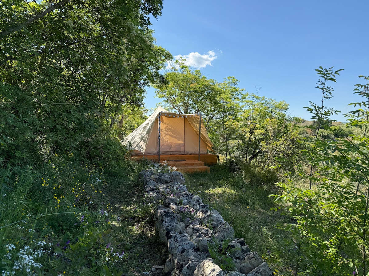 A spacious tent is set amidst lush greenery, partially shaded by leafy trees. Natural stone creates a rustic perimeter, blending with the surrounding landscape. The clear blue sky above contributes to the serene atmosphere of this secluded glamping site.