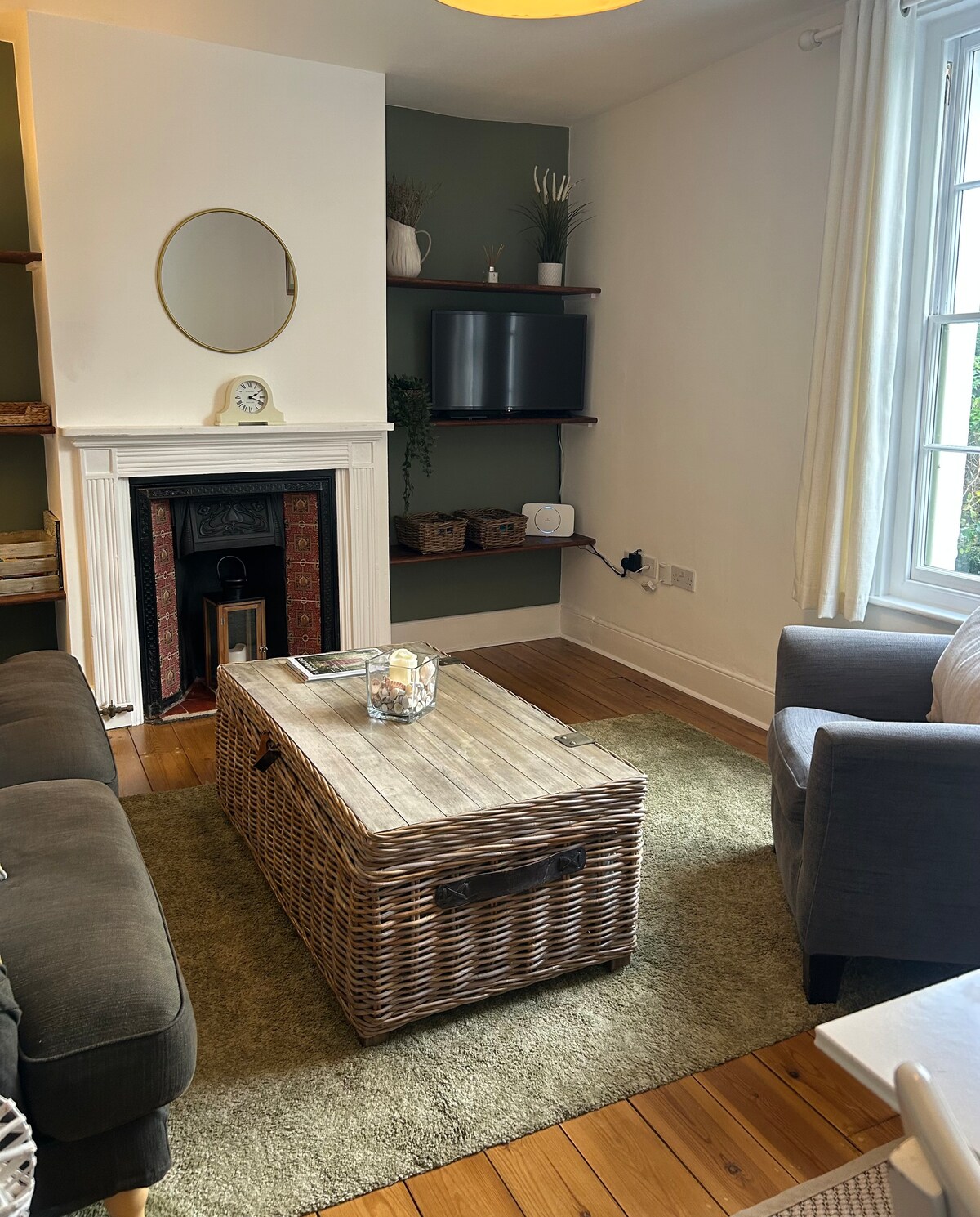 A welcoming living area features a light-colored, textured rug beneath a rectangular, wicker coffee table. A cozy seating arrangement includes a dark sofa and a soft armchair. A decorative mirror hangs above the fireplace, and a flat-screen television rests on a wall-mounted shelf.