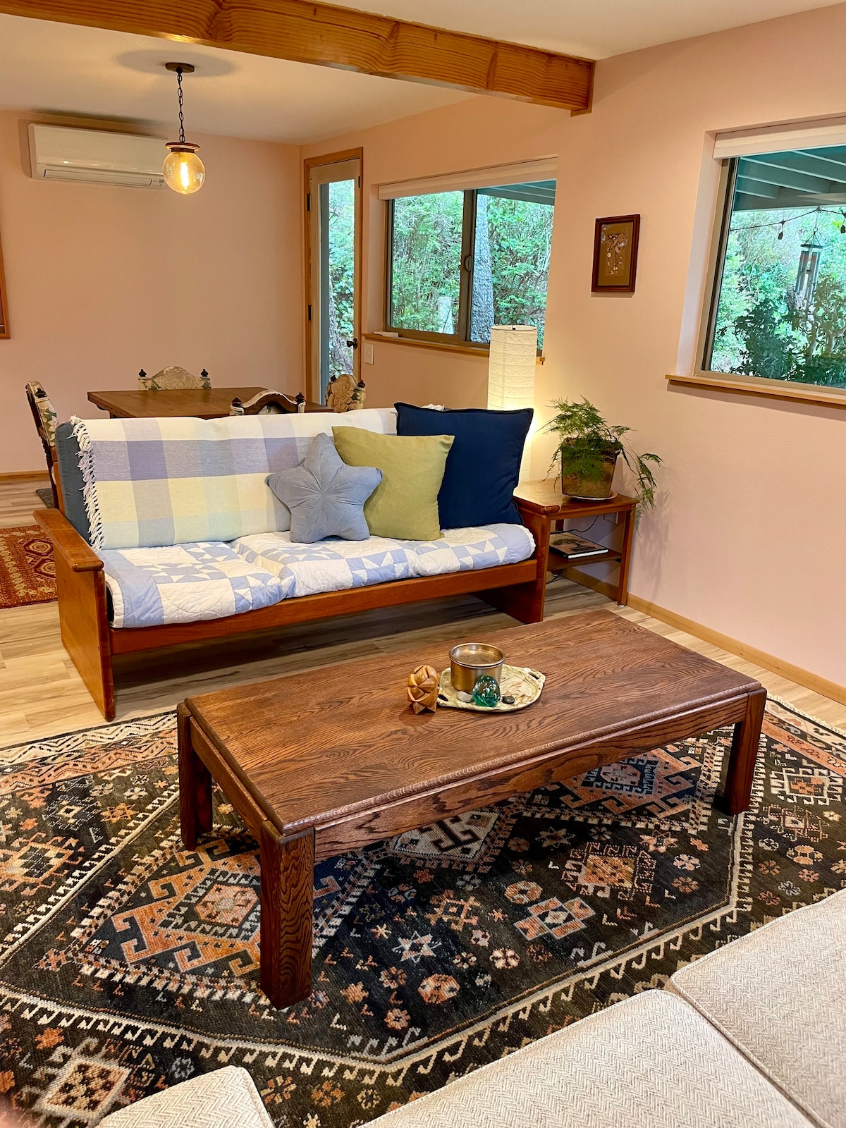A comfortably arranged living area features a light wood coffee table at the center, surrounded by patterned rugs. A small table lamp and a green plant add warmth, while sunlight filters through the nearby windows, revealing light pink walls and a cozy couch adorned with cushions.