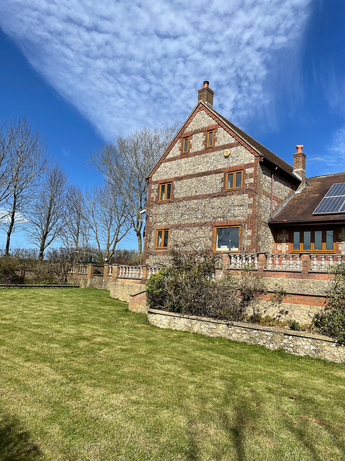 The exterior of the countryside farmhouse is showcased against a blue sky with wispy clouds. A well-maintained lawn leads to the stone structure, featuring large windows framed in warm wood. Surrounding trees provide shade and enhance the natural setting of the property.