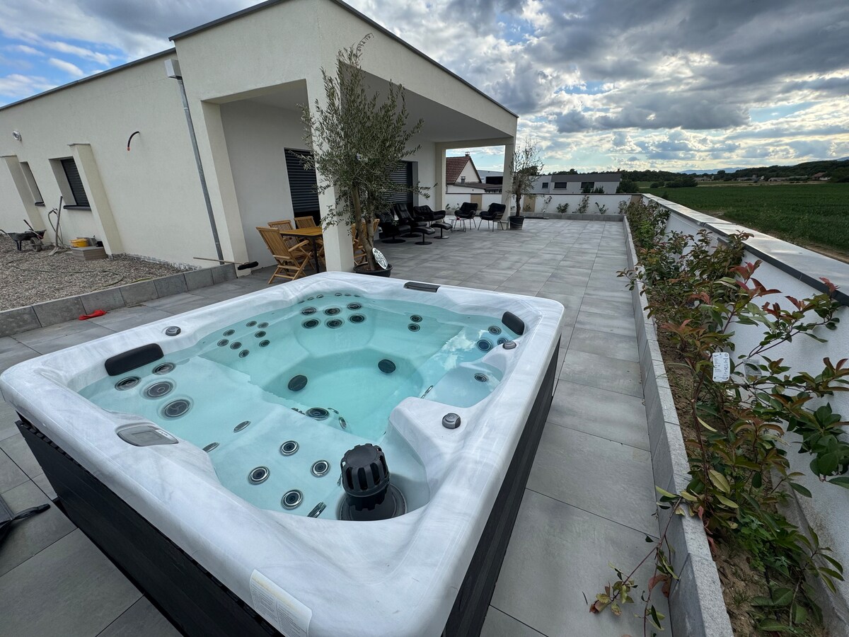 A spacious terrace features a private jacuzzi surrounded by landscaped greenery. Comfortable outdoor seating is arranged nearby under a covered area, offering a relaxing space to enjoy the surroundings. The sky is filled with clouds, adding depth to the tranquil outdoor setting.