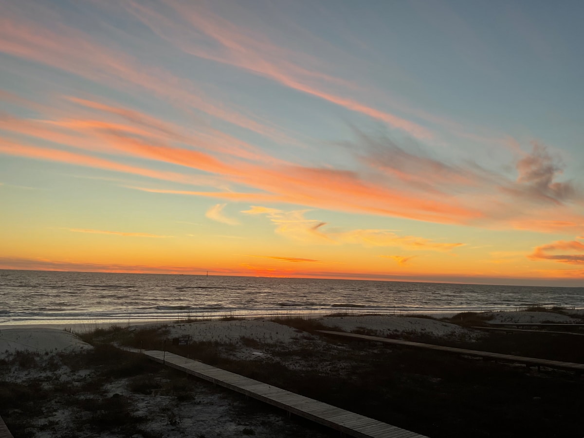 A serene seaside view showcases vibrant hues of orange, pink, and purple in the sunset sky. The tranquil ocean is visible in the foreground, with gentle waves meeting the sandy shore. Boardwalk pathways lead through the beach landscape, enhancing the coastal atmosphere.