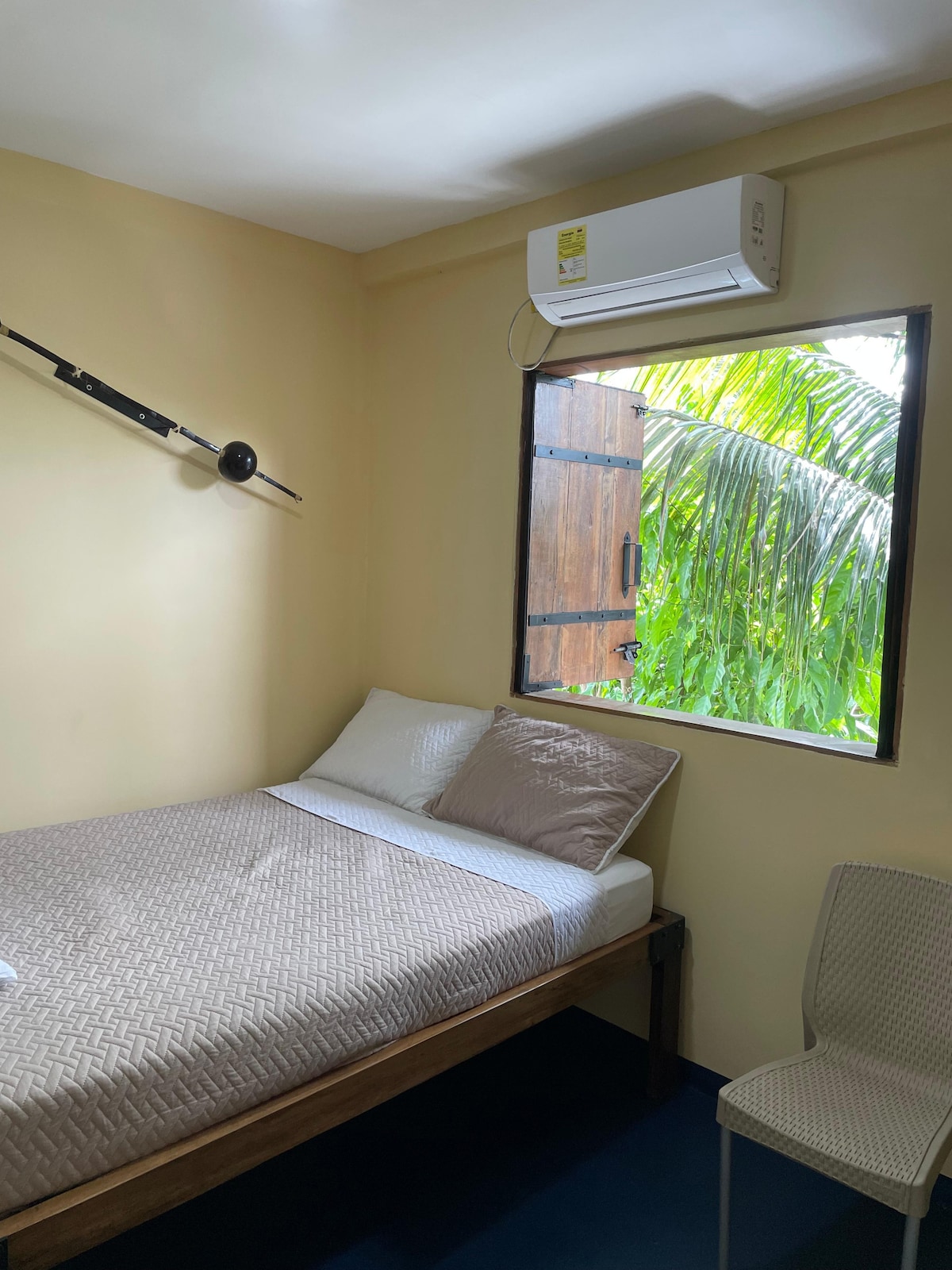 The room features a spacious bed with a beige patterned mattress, positioned against a light yellow wall. A window allows natural light and a view of lush greenery. A wall-mounted air conditioning unit is present, and a simple chair is placed nearby.