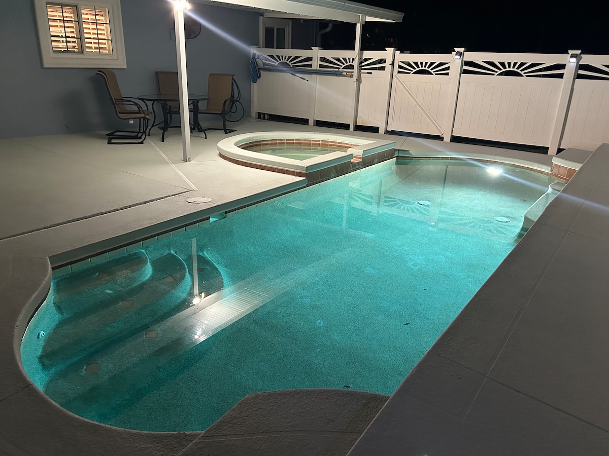 A private pool area is illuminated at night, showcasing a clear water pool and an adjoining jacuzzi. Comfortable seating is visible under a covered patio, offering a relaxed outdoor space. The fence provides privacy while maintaining an inviting atmosphere.