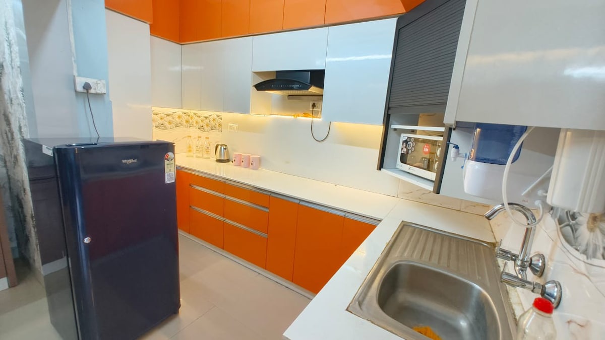 A modern kitchenette features vibrant orange cabinets paired with sleek white surfaces. Essential appliances include a refrigerator, microwave, and kettle. A stainless-steel sink is set into a counter, showcasing a neatly organized space for cooking and meal preparation.