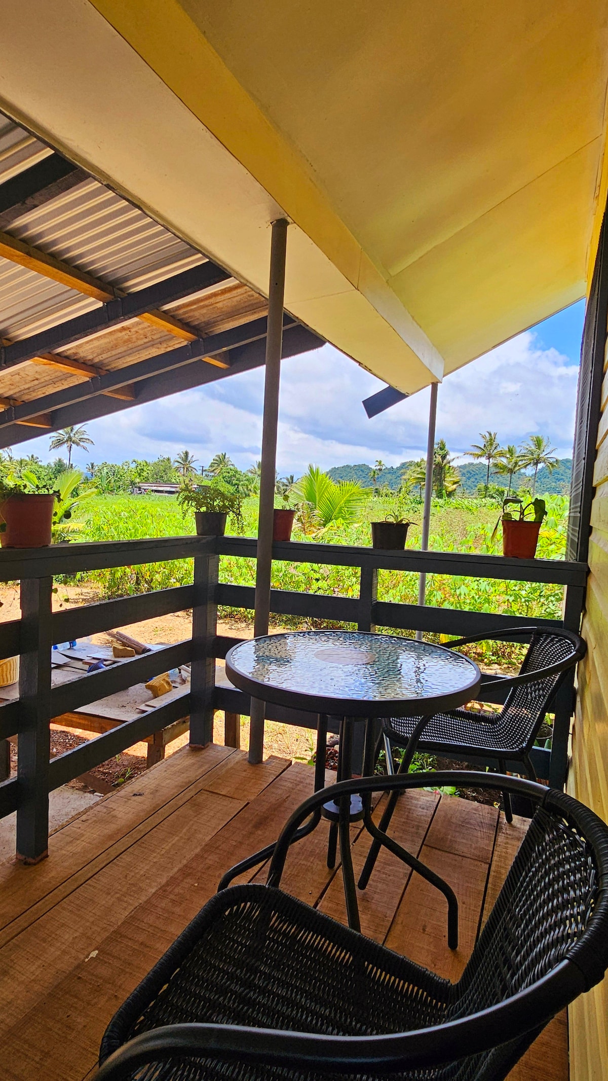 A covered balcony features a circular glass-top table surrounded by two black wicker chairs. Potted plants adorn the railing, offering a view of lush greenery and palm trees in the distance under a bright blue sky.