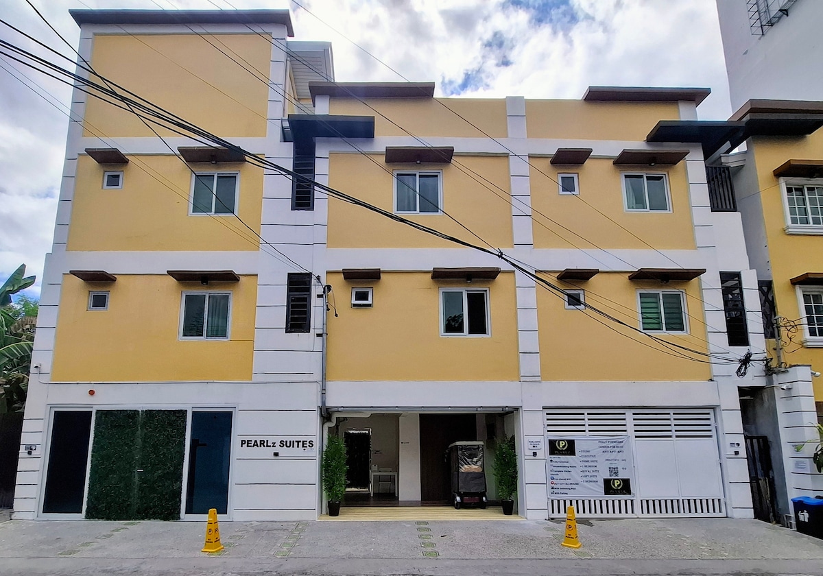 The exterior of PEARLz SUITES Condotel is displayed, featuring a bright yellow facade with multiple windows. A sleek entrance is visible, surrounded by greenery, and a clearly marked sign identifies the establishment. The space includes an accessible parking area, contributing to the convenience of the location.