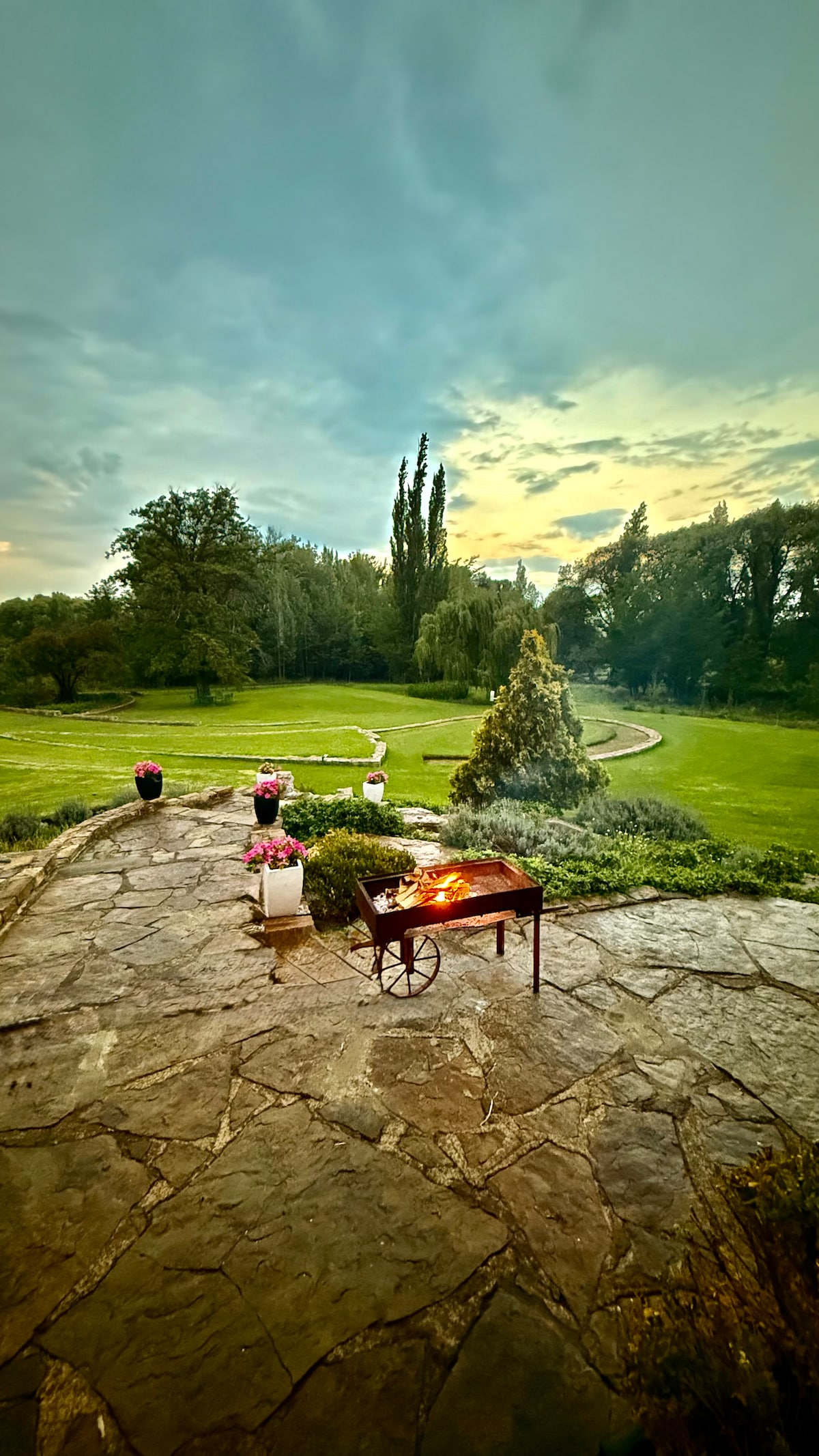 A spacious outdoor area is presented with a stone patio overlooking a well-kept green lawn, surrounded by trees. A charcoal braai is located on the patio, with several pots of colorful flowers positioned nearby. The sky is overcast, hinting at a tranquil evening.