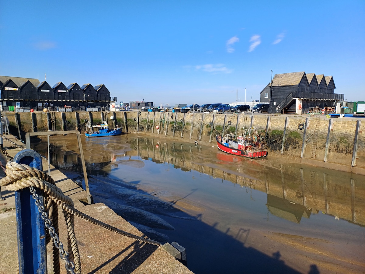 A serene harbour scene features two anchored boats, one in blue and the other in red, resting on the calm water at low tide. The background includes a row of dark wooden buildings and parked vehicles, under a clear blue sky.
