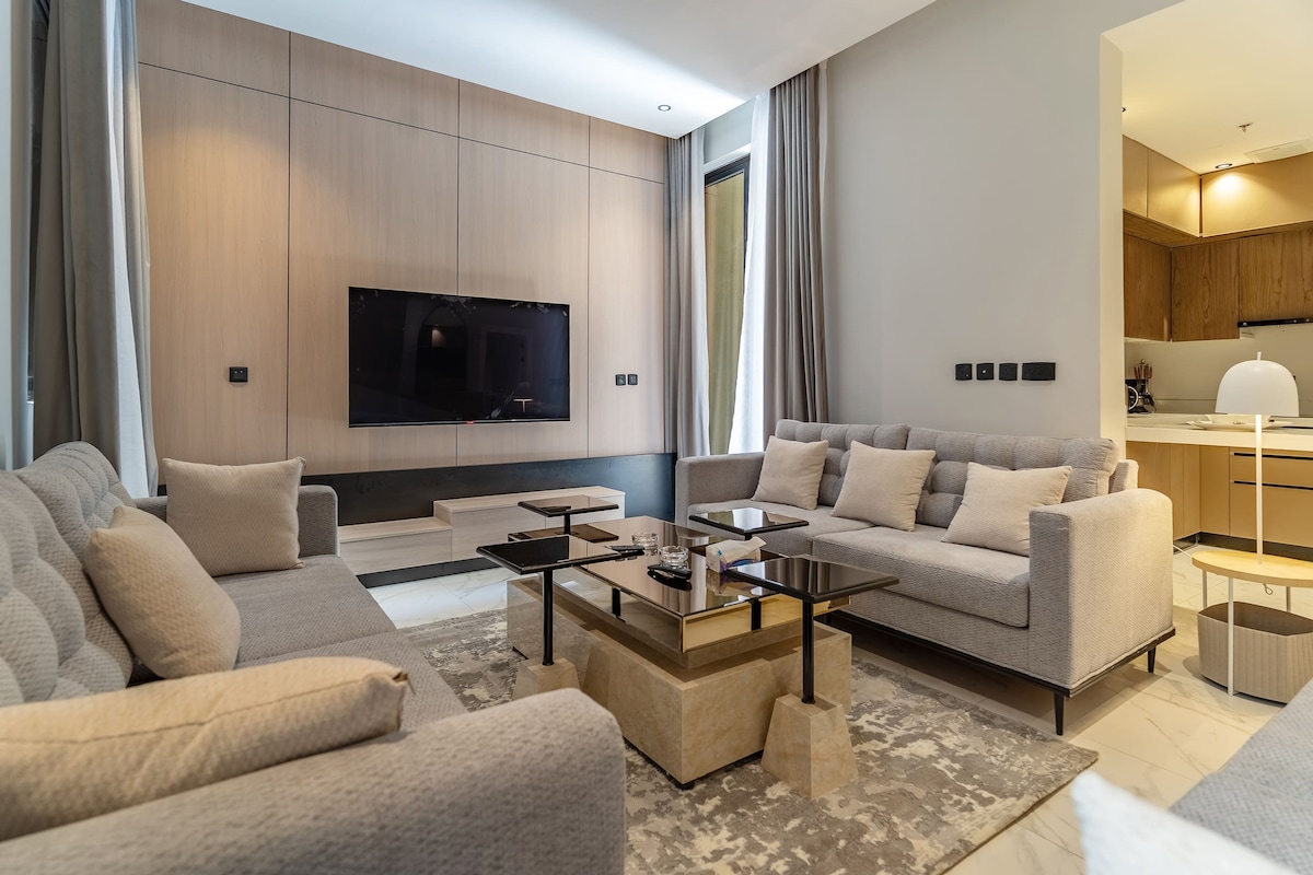 A spacious living area is featured, showcasing soft grey sofas arranged around a sleek glass coffee table. Natural light filters through tall windows, enhancing the simplicity of the neutral-toned decor. A television is mounted on the wall, creating a focal point in the room.