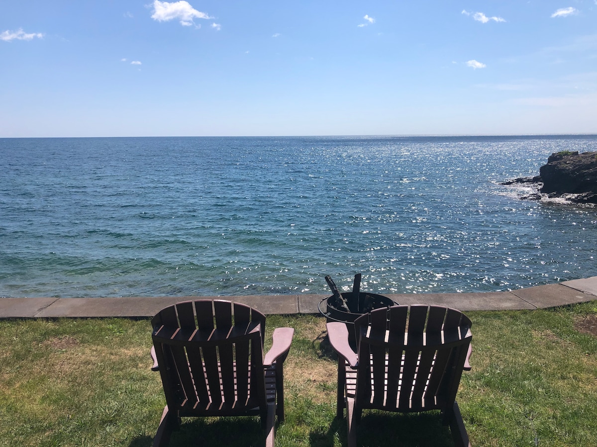 Two wooden adirondack chairs are positioned on the grassy shore of Lake Superior, facing the expansive blue waters. The calm surface reflects the clear sky, with hints of sunlight shimmering on the waves. A fire pit is visible nearby, enhancing the waterfront setting.