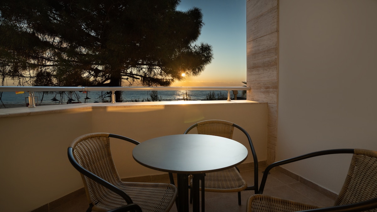 A cozy balcony features a round table accompanied by two woven chairs, facing a scenic view of the ocean and sunset. A tall conifer tree partially frames the space, while the warm glow of the sunset casts soft light across the area.