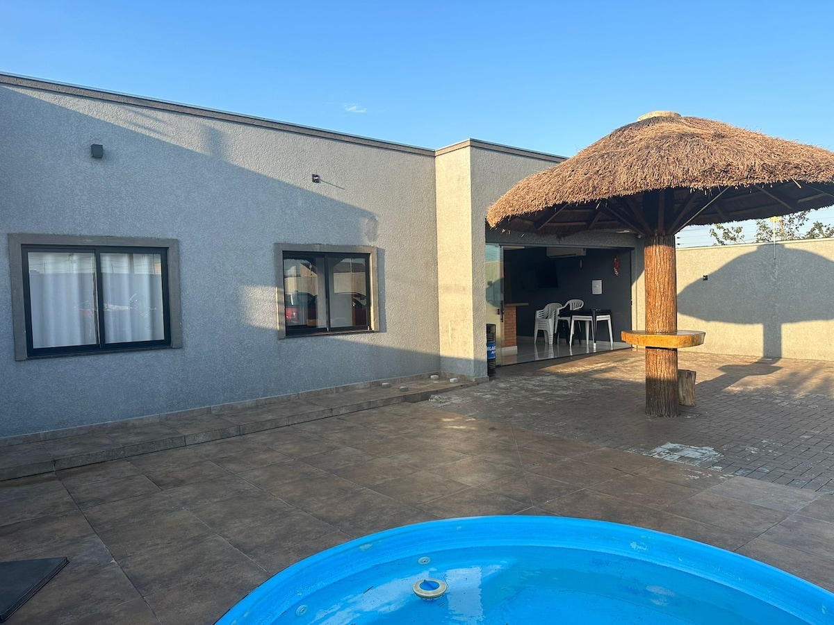 An outdoor area features a blue hot tub in the foreground, with a thatched-roof gazebo nearby. The modern structure is visible in the background, showcasing large windows and a patio area furnished with white chairs, providing a spacious setting for relaxation.