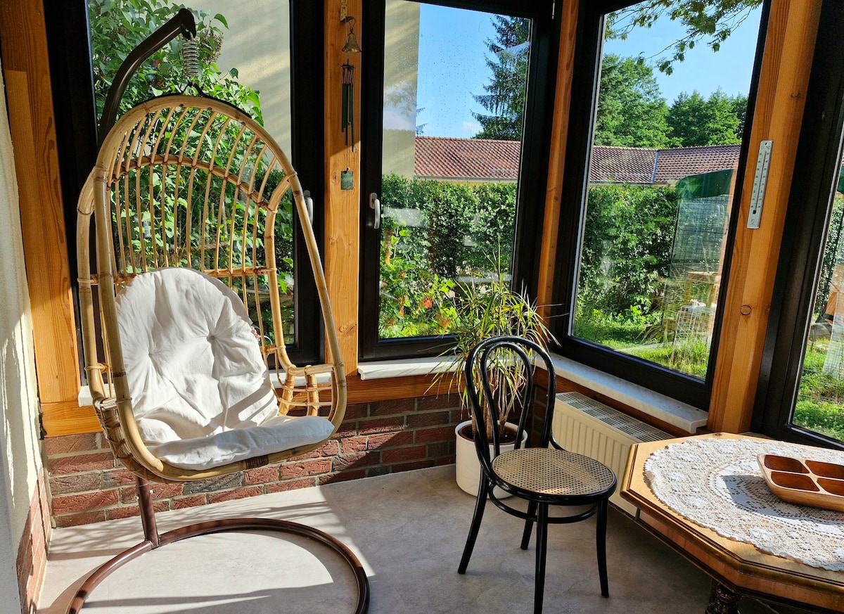 A bright winter garden area is depicted, featuring a hanging rattan chair with a soft cushion, a simple chair beside it, and a table with a decorative lace tablecloth. Large windows offer views of the lush garden outside, enhancing the open atmosphere.