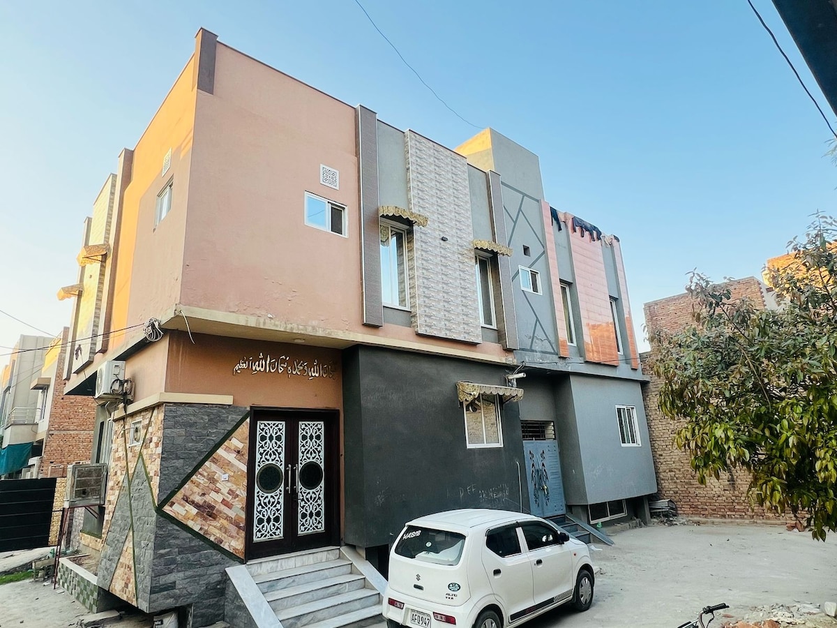 The exterior of the three-bedroom house is shown, featuring a mix of colors and textures on the façade. Distinct architectural details are visible, including decorative elements around the entrance and large windows. A compact white car is parked in front, amidst a spacious driveway.